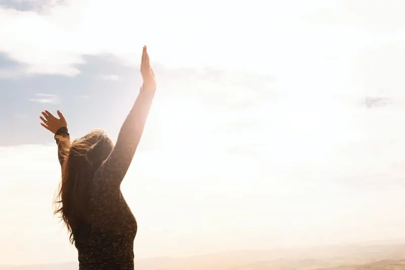 woman raising both hands in the air facing a very white light and clouds