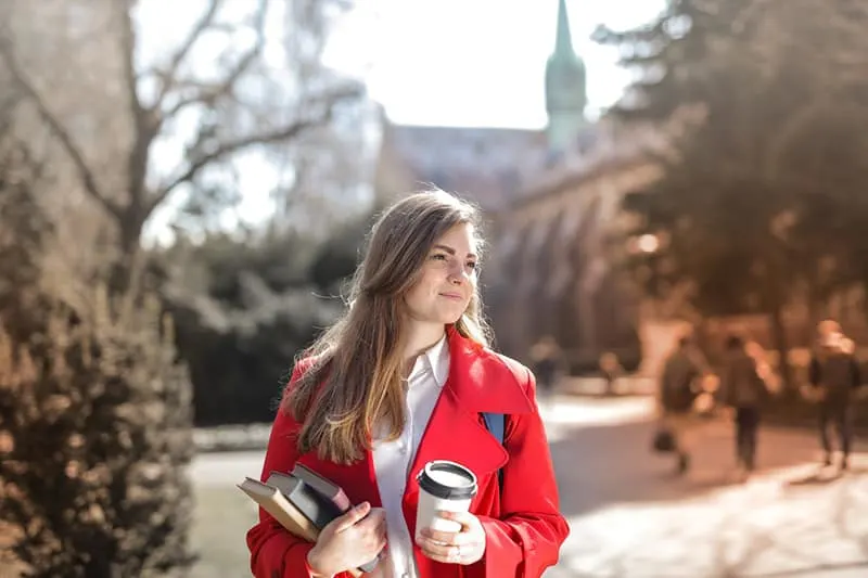 Woman wearing red coat and holding her books and a coffee cup