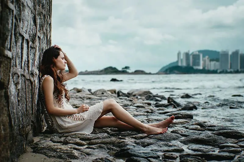woman sitting in the rocks beside a body of the water leaning a wall