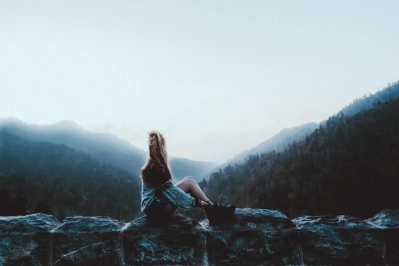 woman sitting on gray rock facing green mountains