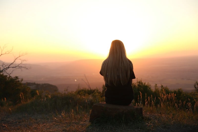 blonde woman sitting on rock and looking at sunset