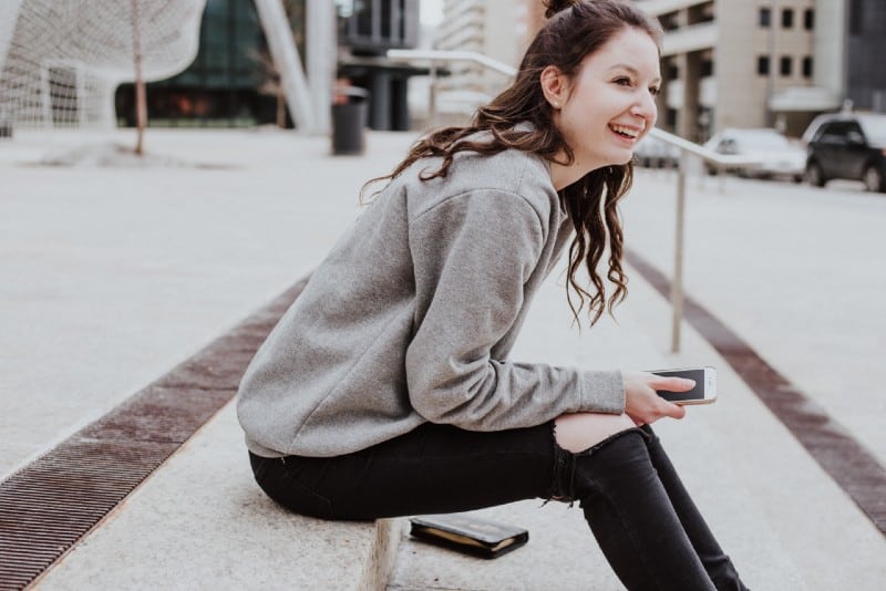 mulher sentada no passeio com um smartphone na mão e a sorrir 