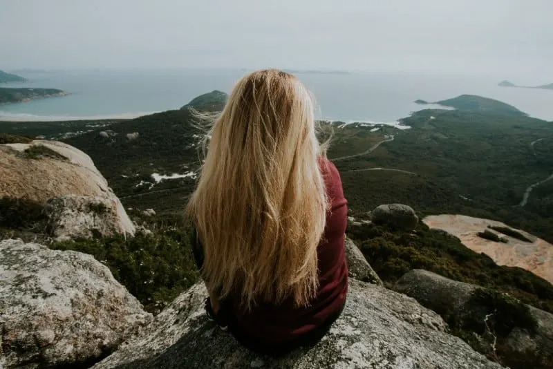 blonde woman sitting on top of hill looking at sea