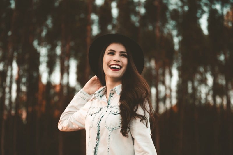 mulher sorridente com chapéu preto numa floresta