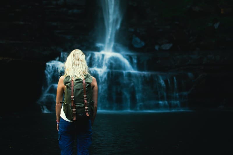 blonde woman with backpack standing near water falls