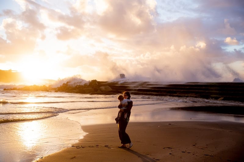 mulher à beira-mar com um bebé ao colo durante o pôr do sol