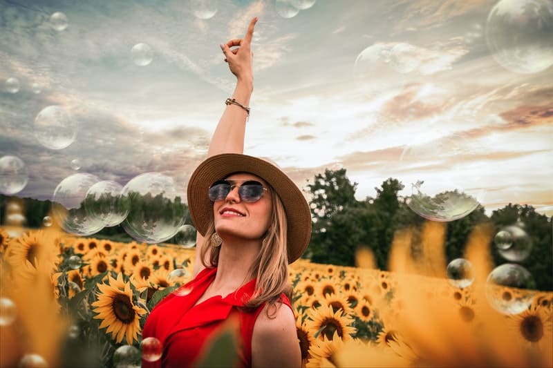 woman surrounded by sunflowers with one hand raised wearing hat and sunglasses