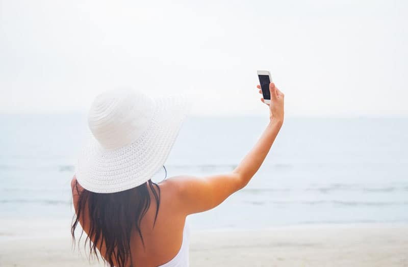 donna che scatta una foto indossando un abito estivo con cappello bianco in piedi vicino allo specchio d'acqua
