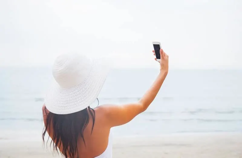 woman taking a picture wearing a summer suit with white hat standing near the body of water