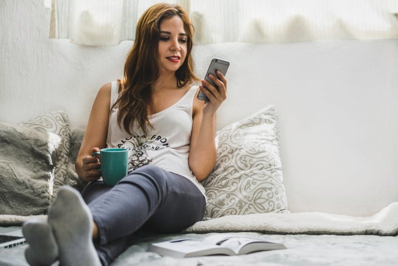 woman in white top holding mug and using smartphone