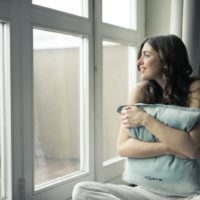 mujer de pelo negro abrazando una almohada gris cerca de una ventana de cristal