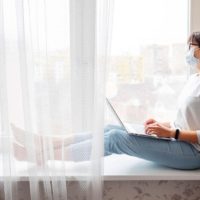 donna in autoisolamento che indossa una maschera facciale mentre è seduta sul vetro di una finestra con un computer portatile in grembo