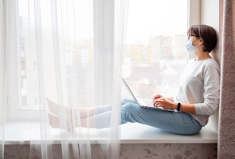woman on self isolation wearing face mask while sitting on window pane laptop on her lap