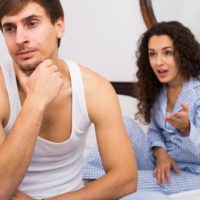 woman in pajamas nagging a man in white tank top while sitting on bed