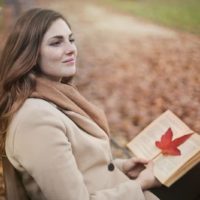 mujer de pelo rojo sonriendo sentada en un parque sosteniendo un libro con una hoja en él durante la temporada de otoño