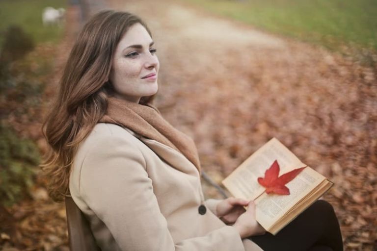 red haired woman smiling sitting on a park holding a book with a leaf in it during autumn season