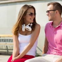 woman in white top and red pants with headphone round her neck sitting next to a guy in pink shirt sitting outside in daytime