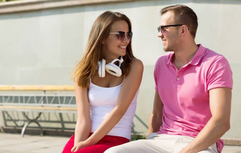 donna in top bianco e pantaloni rossi con cuffie al collo seduta accanto a un ragazzo in camicia rosa seduto all'aperto di giorno