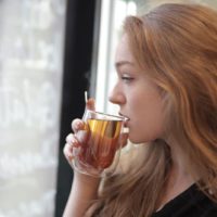 mulher a beber chá de um copo de chá enquanto olha para o exterior através de uma janela de vidro