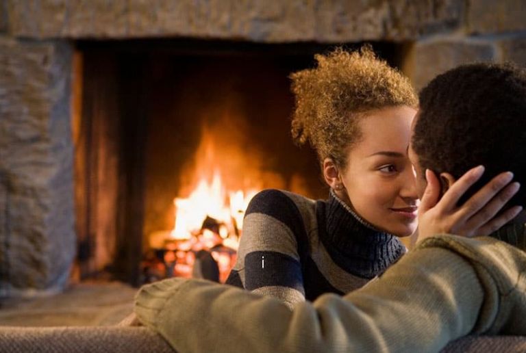 woman touching the face of a man sitting in a couch in front of the fireplace