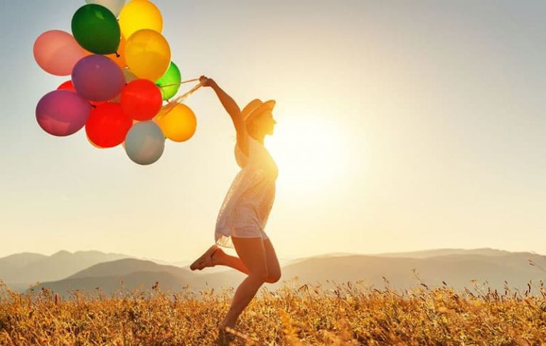 happy woman hopping in the middle of the field carrying different colored balloons