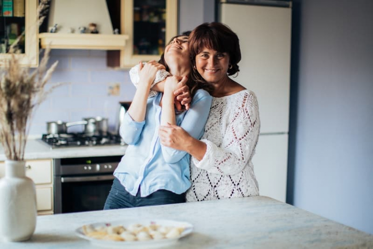 woman in white knitted lace dress hugging a woman laughing inside the kitchen