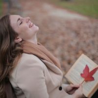 mujer alegre sentada en un banco sonriendo y sintiéndose feliz mientras sostiene un libro con una hoja roja
