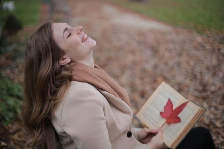 joyful woman sitting on a bench smiling and feeling happy while holding a book with a red leaf