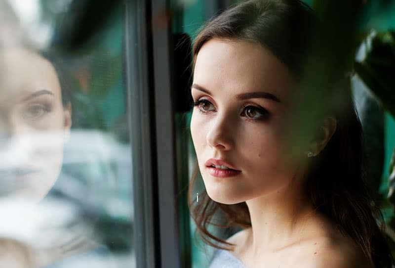 focus photography of a woman near glass window with green leaves nearby