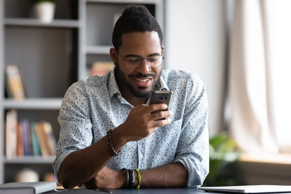 a smiling black man sits in the office and writes an sms