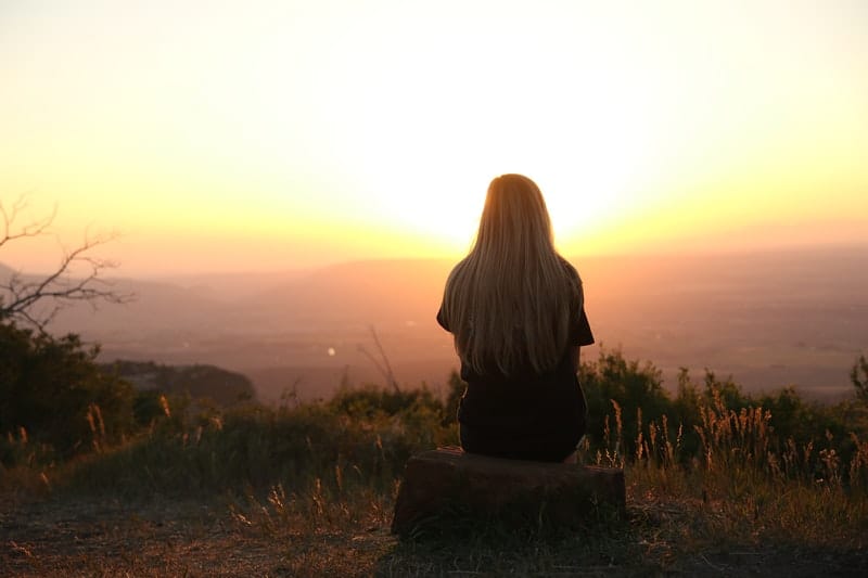 dietro di donna che guarda il sole al tramonto mentre è seduta su una roccia in cima alla montagna