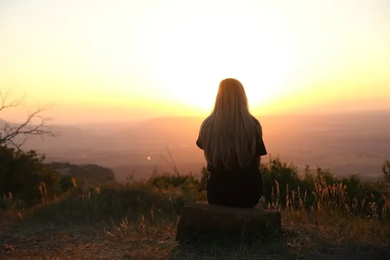behind of woman looking at the setting sun while sitting on a rock at the top of the mountain