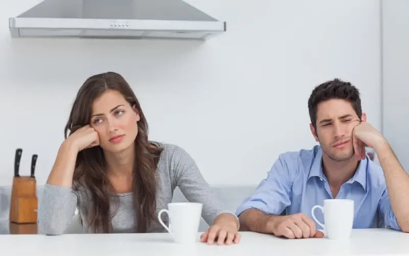 Bored couple sitting at table next to each other with white mugs in front of them