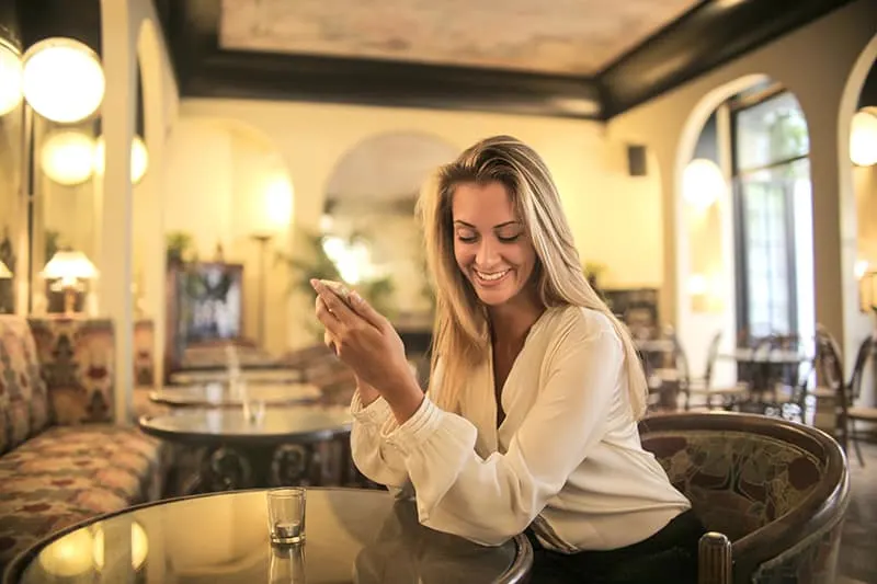 cheerful female having drink in elegant bar while using smartphone