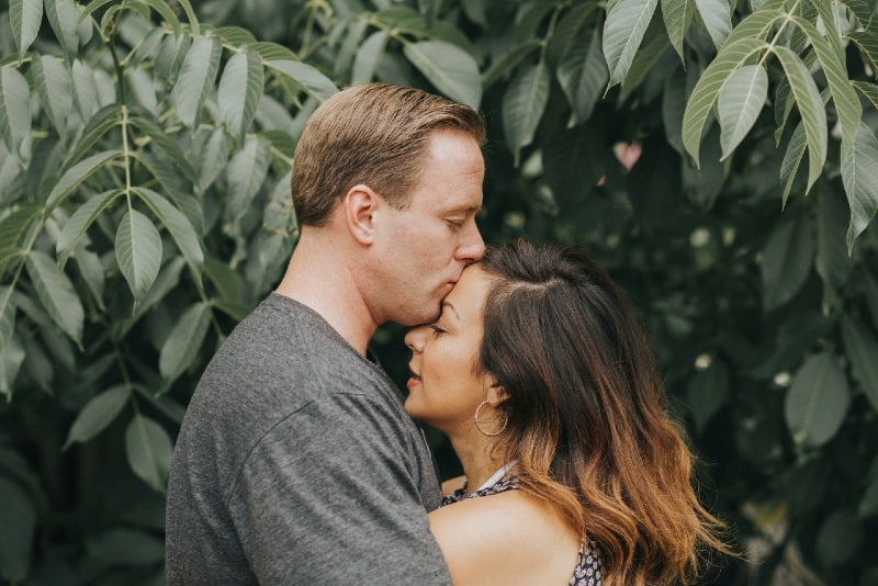 man hugging and kissing woman on forehead near tree leaves