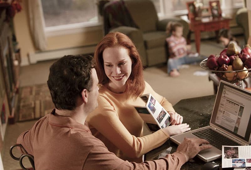 couple planning a vacation looking at each other with a laptop on table and woman holding pictures and a kid playing at a distance