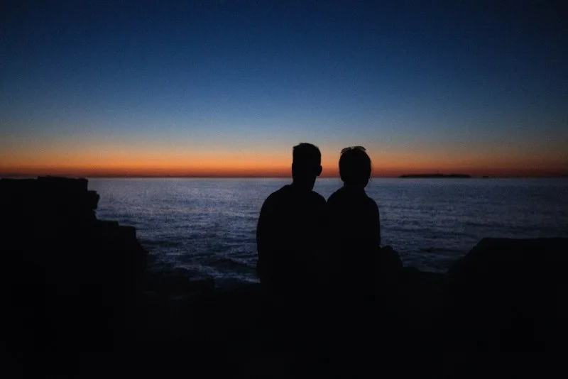 man and woman sitting on rock looking at sea