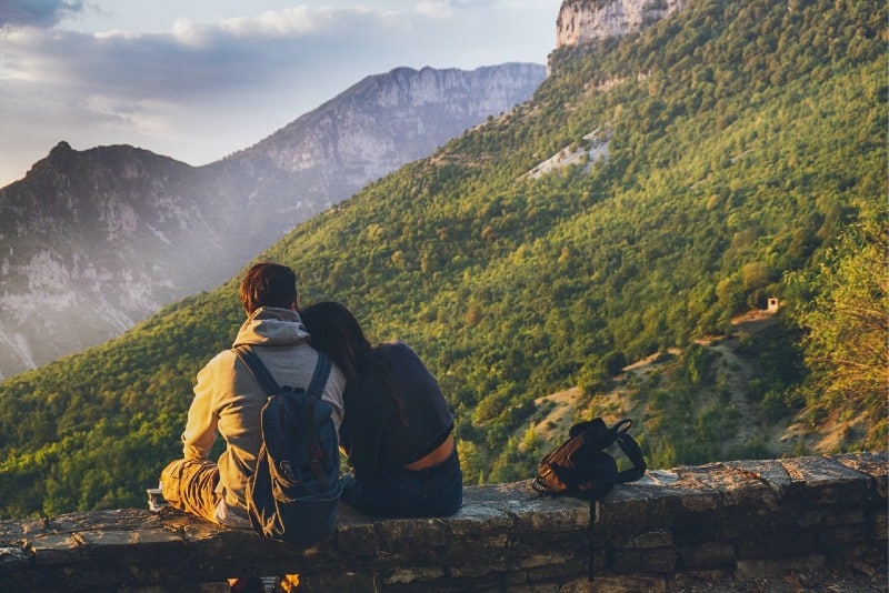 man with backpack and woman sitting outdoor looking at mountain