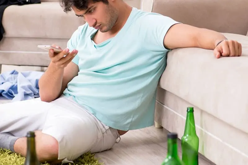 drunk man texting while sitting on the floor leaning on sofa inside living room with empty bottles around