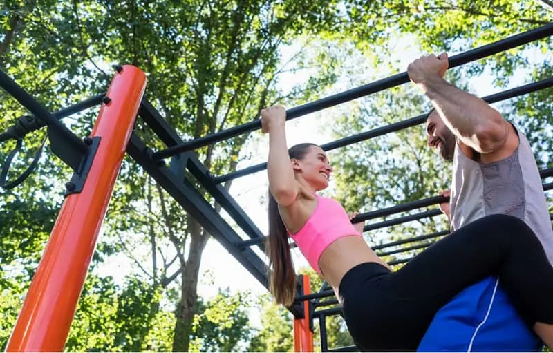 fit young woman smiling while doing pull ups supported by her partner during couple workout