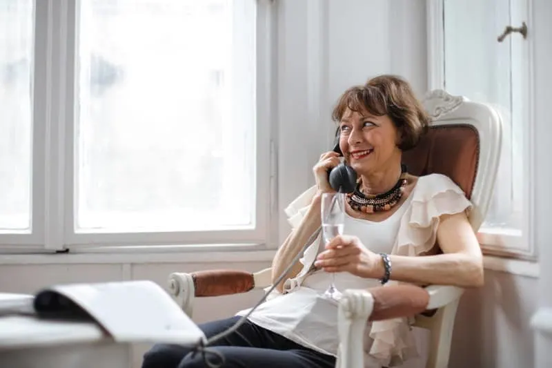 happy middle aged woman making phone call while holding a glass and sitting near the window