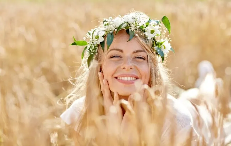Happy beautiful blonde woman wearing wreath of flowers lying on the ground in a cereal field