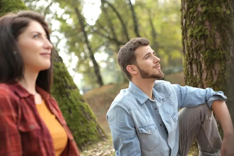 man and woman thoughtfully looking away while sitting near trees in the park