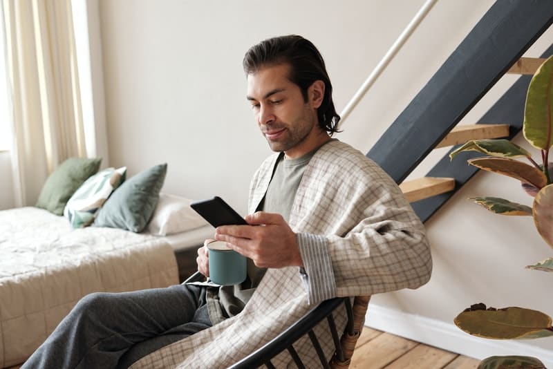 uomo che controlla il cellulare mentre prende il caffè e si siede vicino al letto in accappatoio