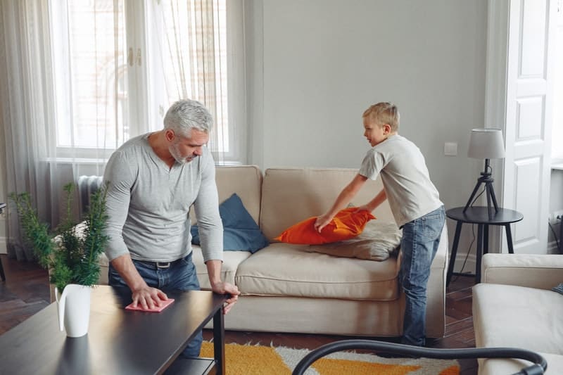 uomo che pulisce il tavolo e un bambino che tiene in mano il cuscino arancione