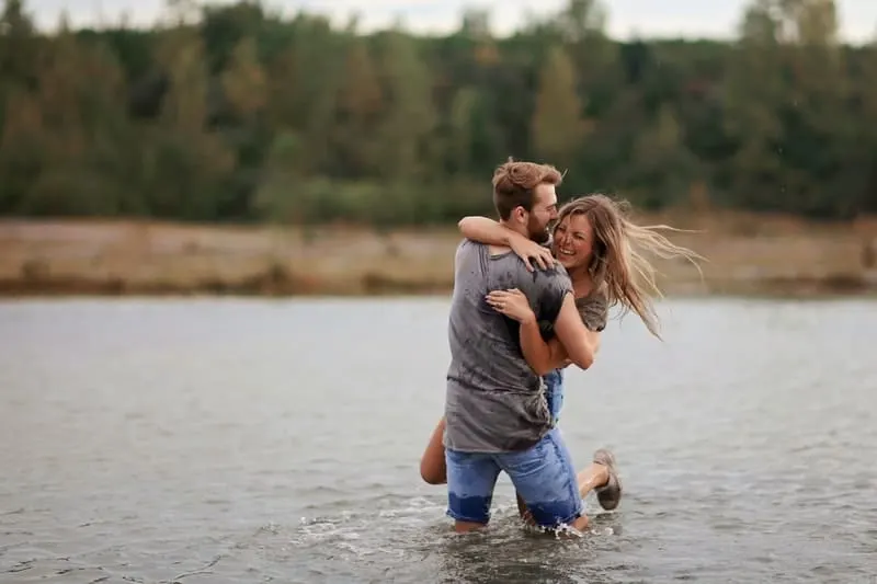 man hugging laughing woman while standing in the middle of a body of water