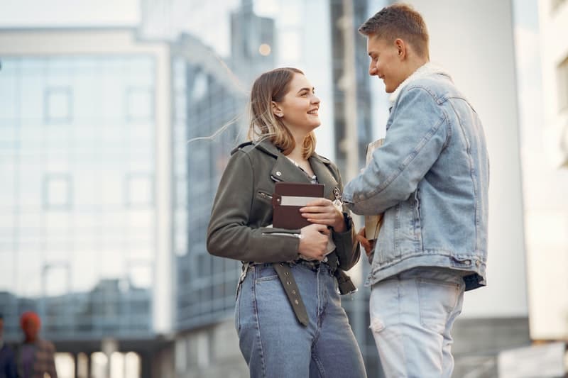 man in blue denim jacket and woman in gray coat talking somewhere outside a building