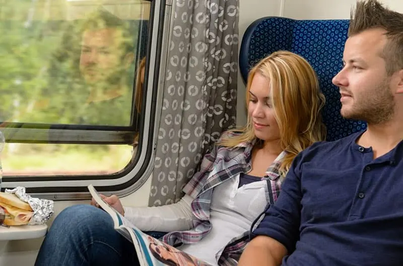 man looking outside the train's window with a woman beside her reading magazine
