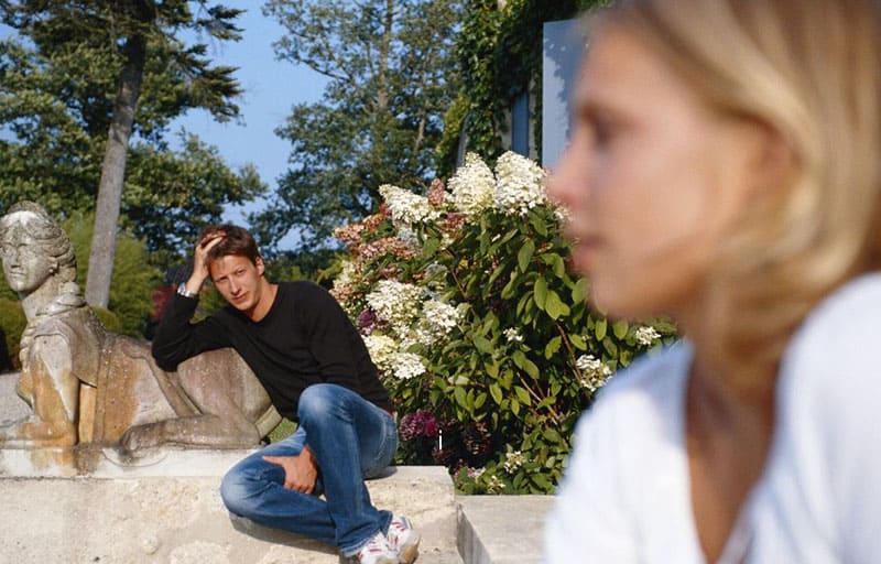man sitting near the garden staring at a woman in blurred distant from him