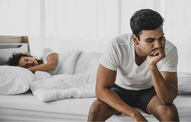 man thinking near his wife sleeping in the same bed he is sitting